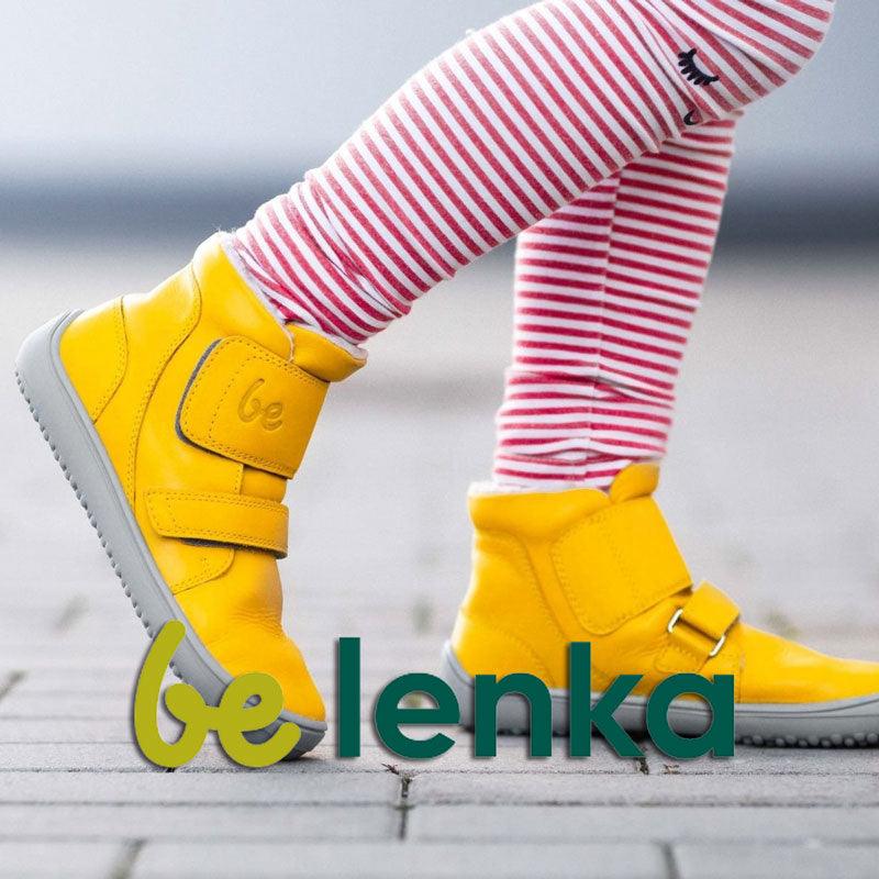 Ett barn leker i ett par gula barfotaskor från varumärket Be Lenka. Skorna är inzoomade nära och man ser även att strumpbyxorna är randiga i rött och vitt.