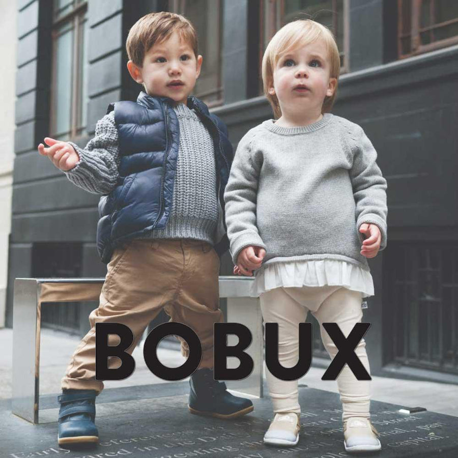 Två små pojkar står i en stadsmiljö i fina skor från Bobux och är nyfikna på vad som händer.