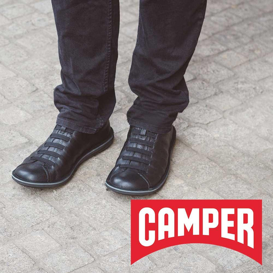 Ett barns fötter i ett par barfotaskor i svart från märket Camper står på ett torg.