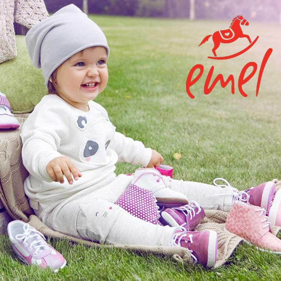 Ett litet barn sitter på en äng under sommaren och har massor av barnskor framför sig från barnskomärket Emel.