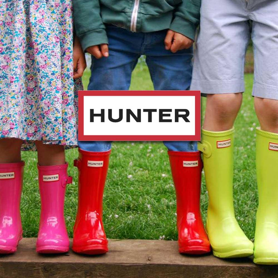 Tre barn står i blött gräs med färgglada gummistövlar från märket Hunter.