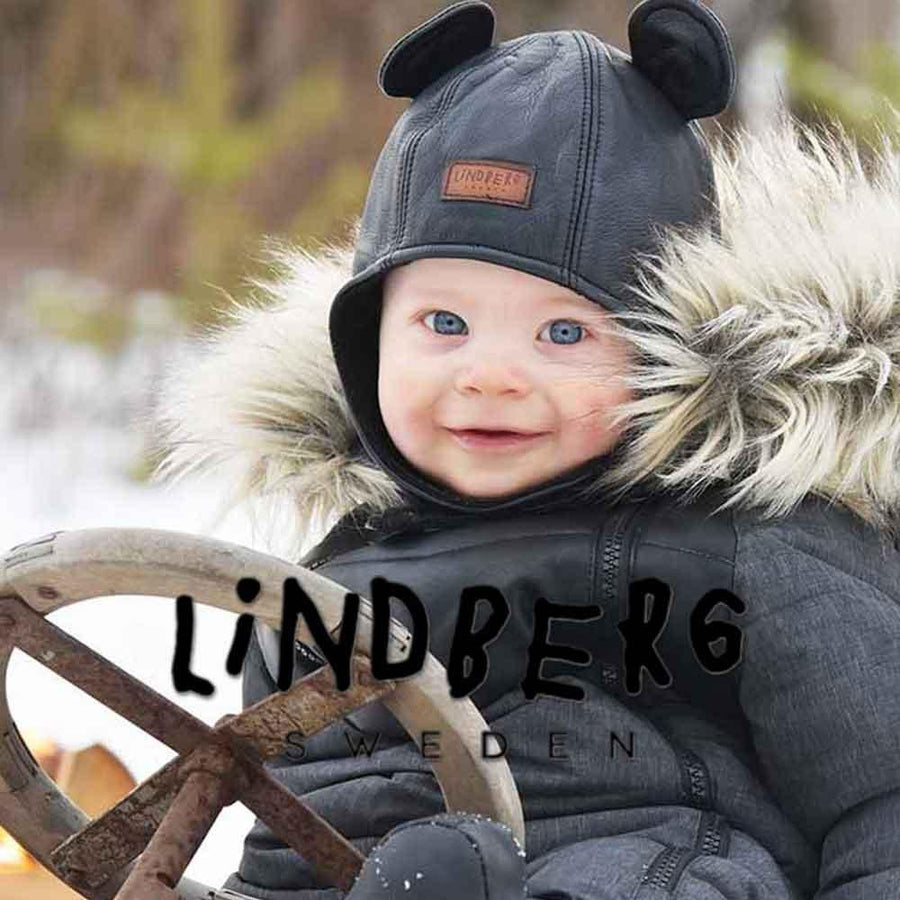 En glad liten unge ler mot kameran medan den sitter på en pulka under vintern i kläder från märket Lindberg.