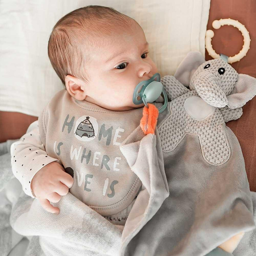 Ett litet barn, en bebis, ligger i sin säng med en napp i munnen som är fäst med en napphållare till filten.
