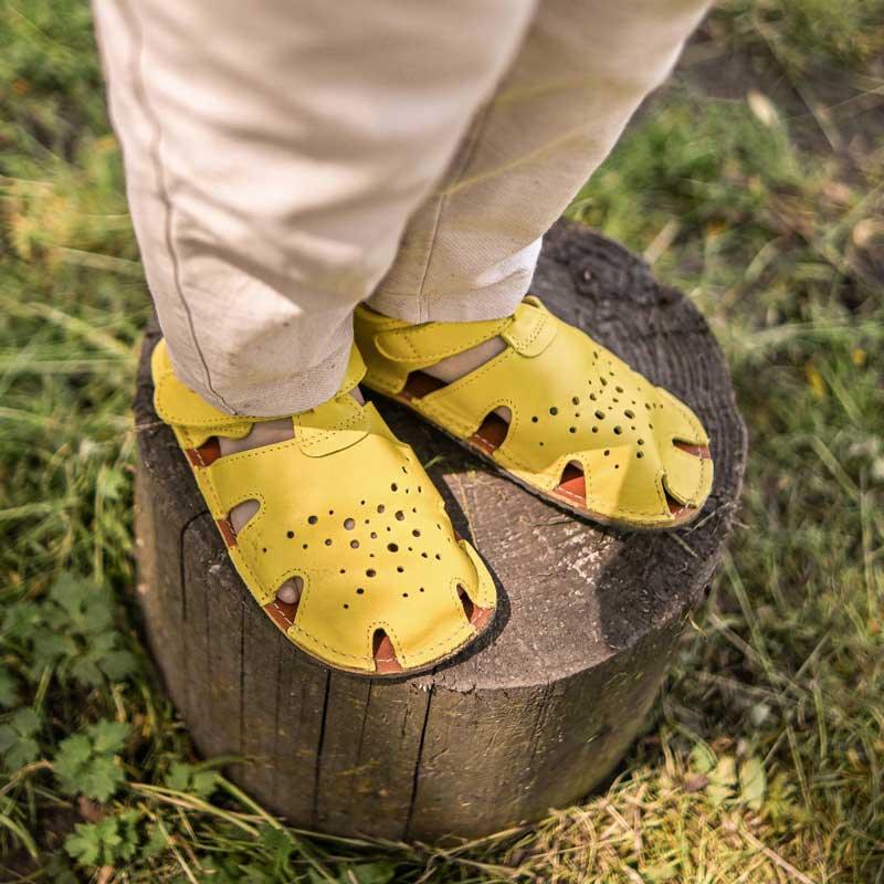 Ett barn står på en stubbe i ett par gula barfotaskor från märket Tikki.