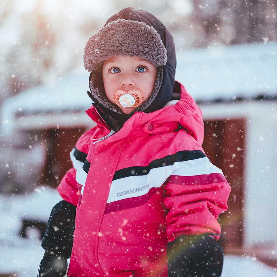 Ett barn iklädd en varm rosa vinteroverall står utomhus medan det snöar och har en napp i munnen.