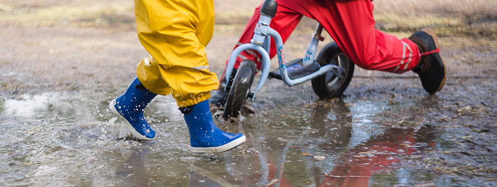Två barn leker i vattenpölar med färgglada gummistövlar på fötterna.